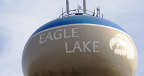 A tractor plows a sunlit field at dawn, creating dust while lush green crops flourish along the edge of the dirt path