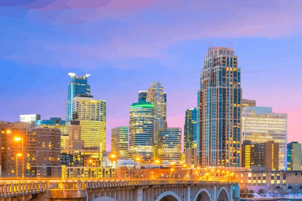 A vibrant twilight cityscape showing glowing high-rise office buildings and a lit bridge in the foreground against a colorful sunset sky.